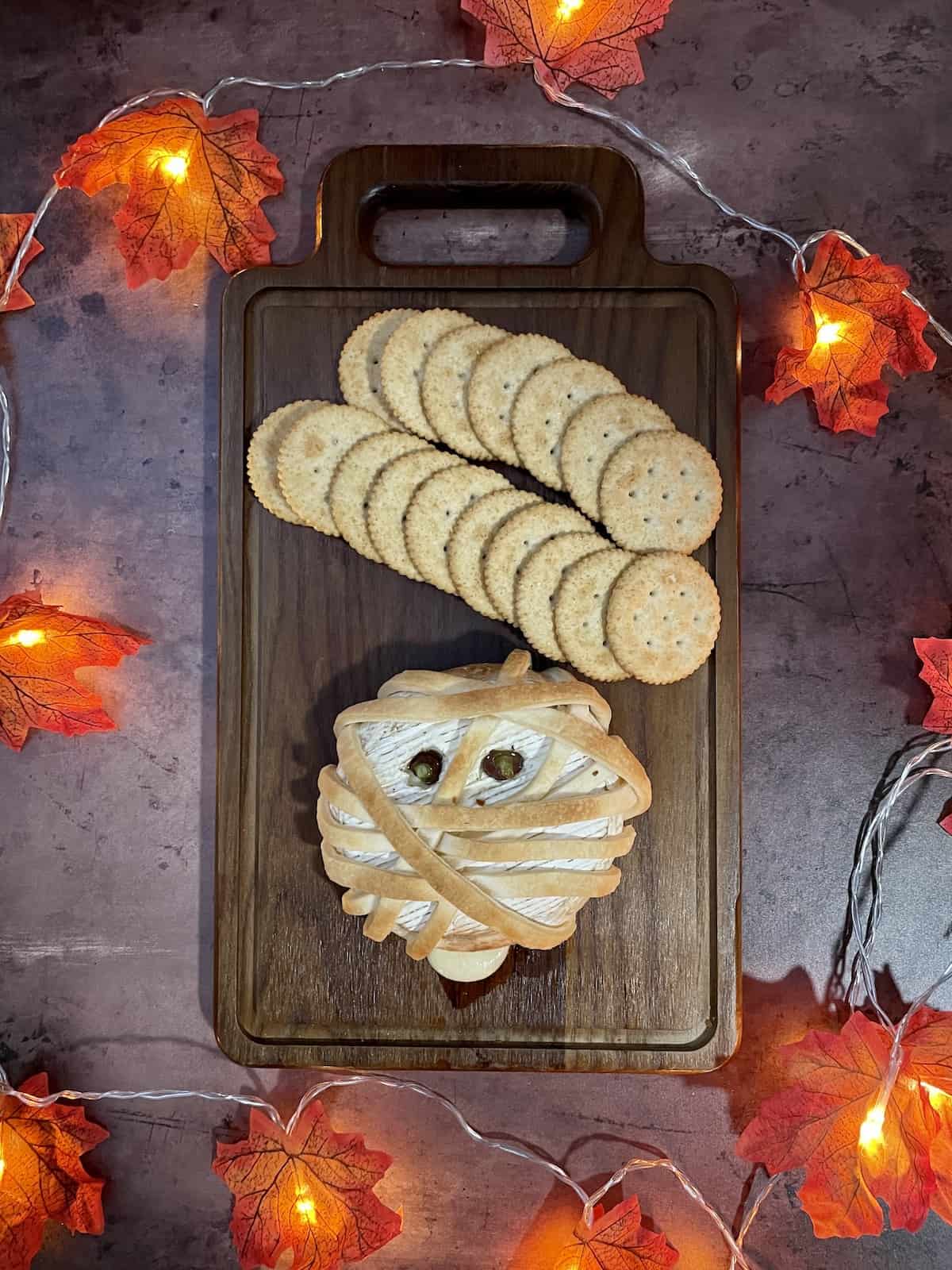 Halloween Mummy dough wrapped brie on wood board with lighted fall leaves around it.