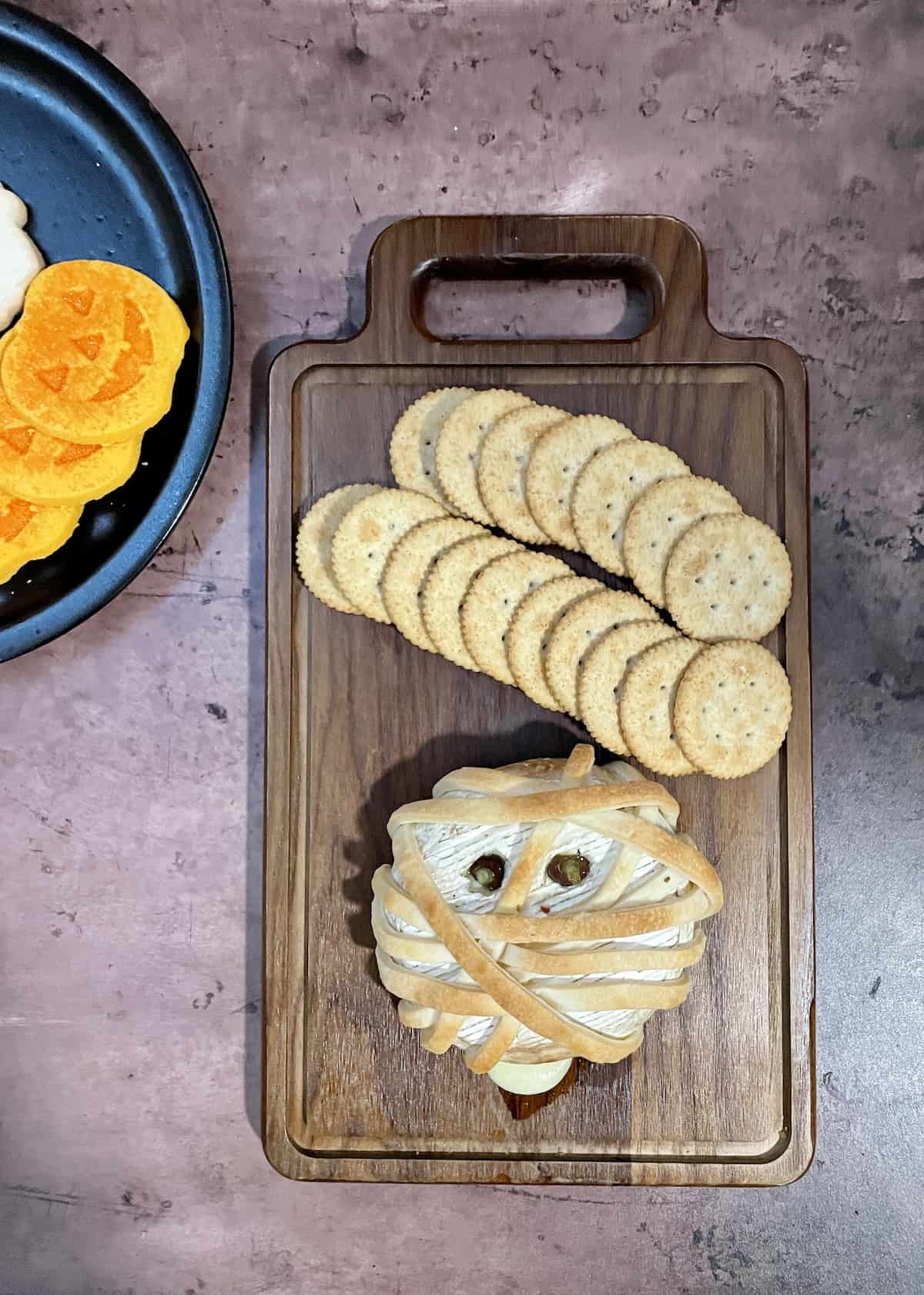 Brie cheese with dough strips wrapped around to look like mummy face with crackers on wood cutting board.