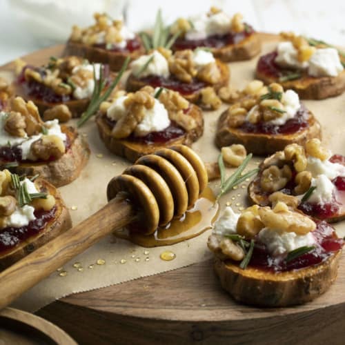 Sweet potato slices topped with cranberries, walnuts, goat cheese, honey, and rosemary on a wood slab.