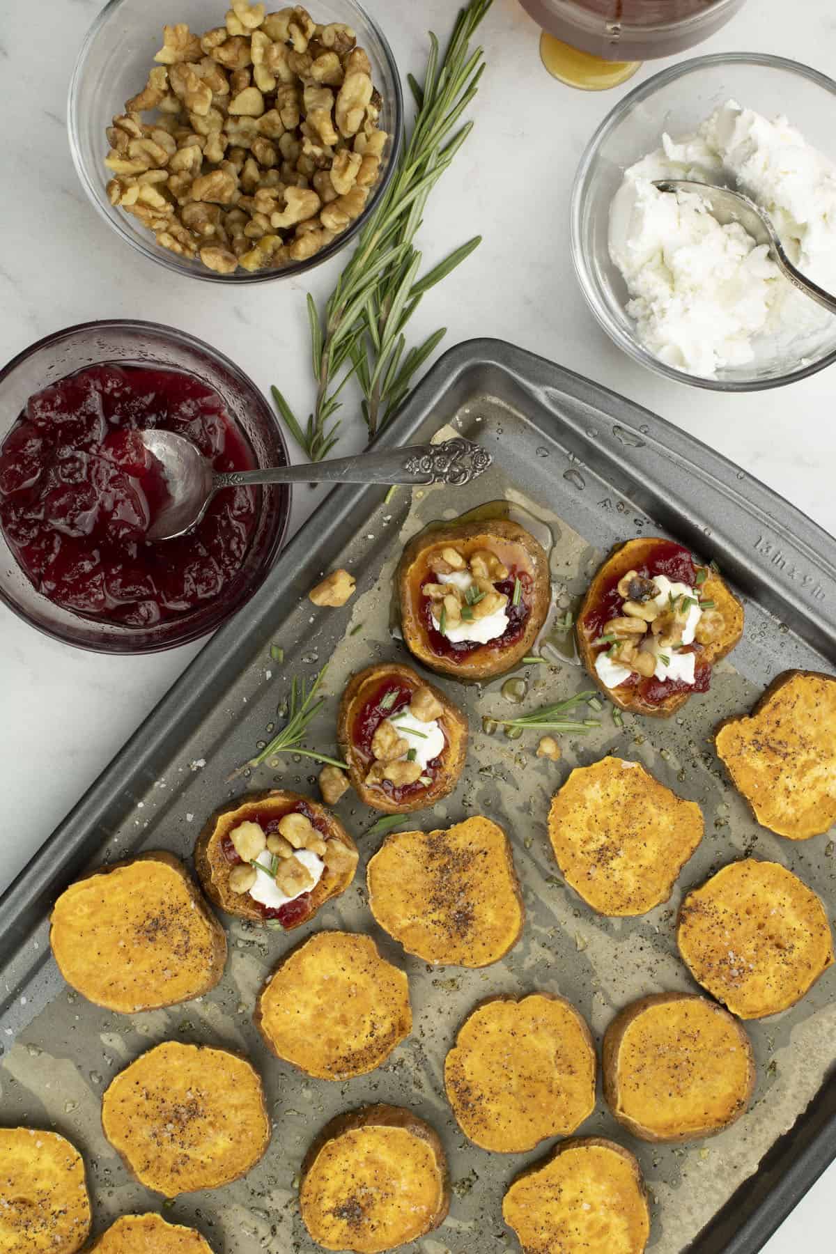 Sweet potato slices topped with cranberries, walnuts, goat cheese, honey, and rosemary on a baking sheet.
