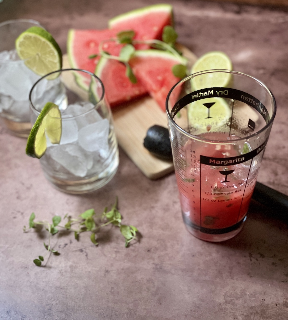 Cocktail shaker with mint and watermelon, glass with ice, cutting board with watermelon and lime.