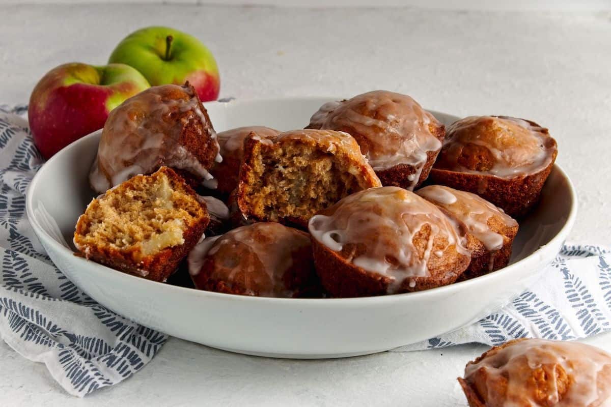 Apple fritters with icing in a white bowl on a white table.