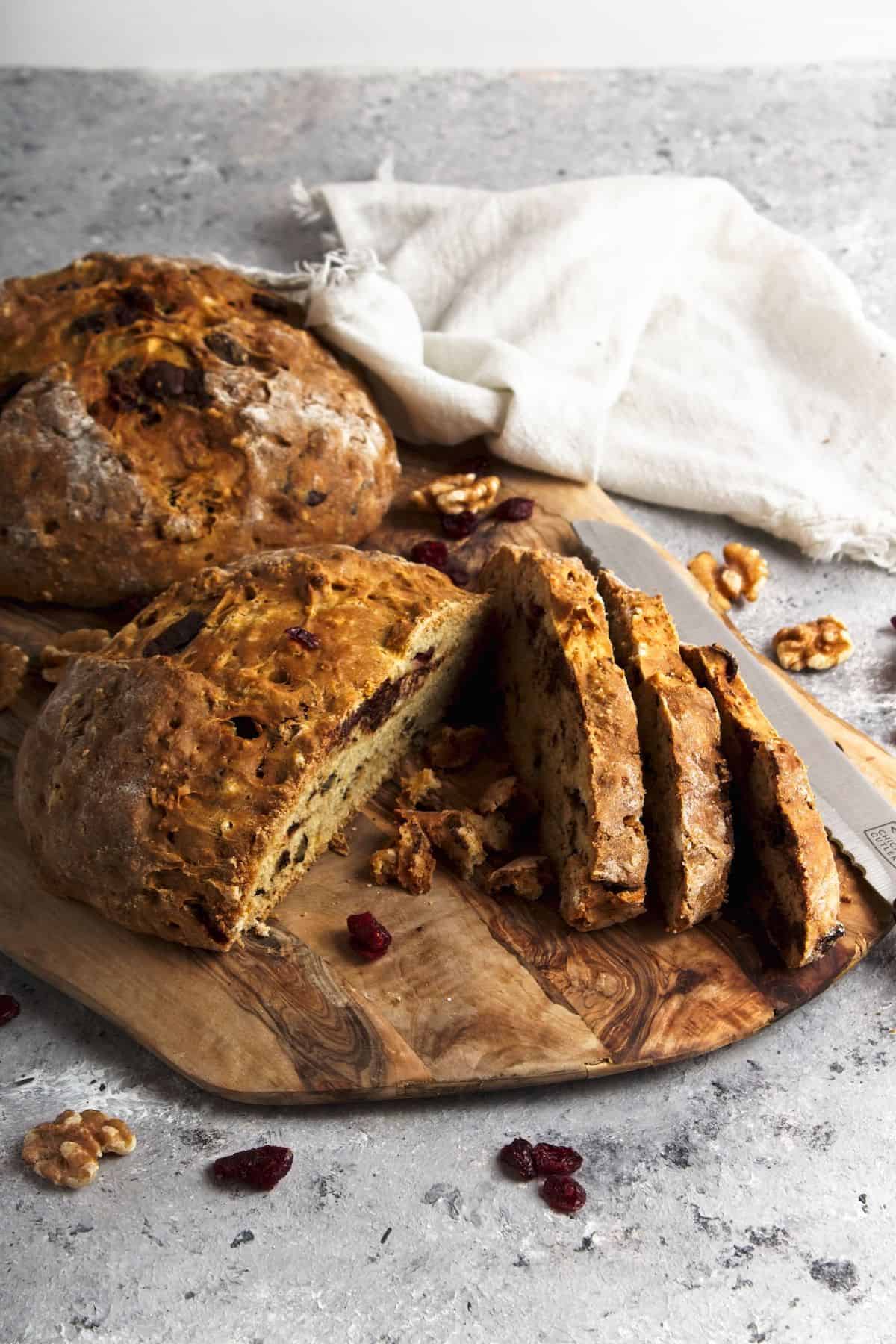 Bread with cranberries, walnuts, and chocolate chips partially sliced on cutting board.