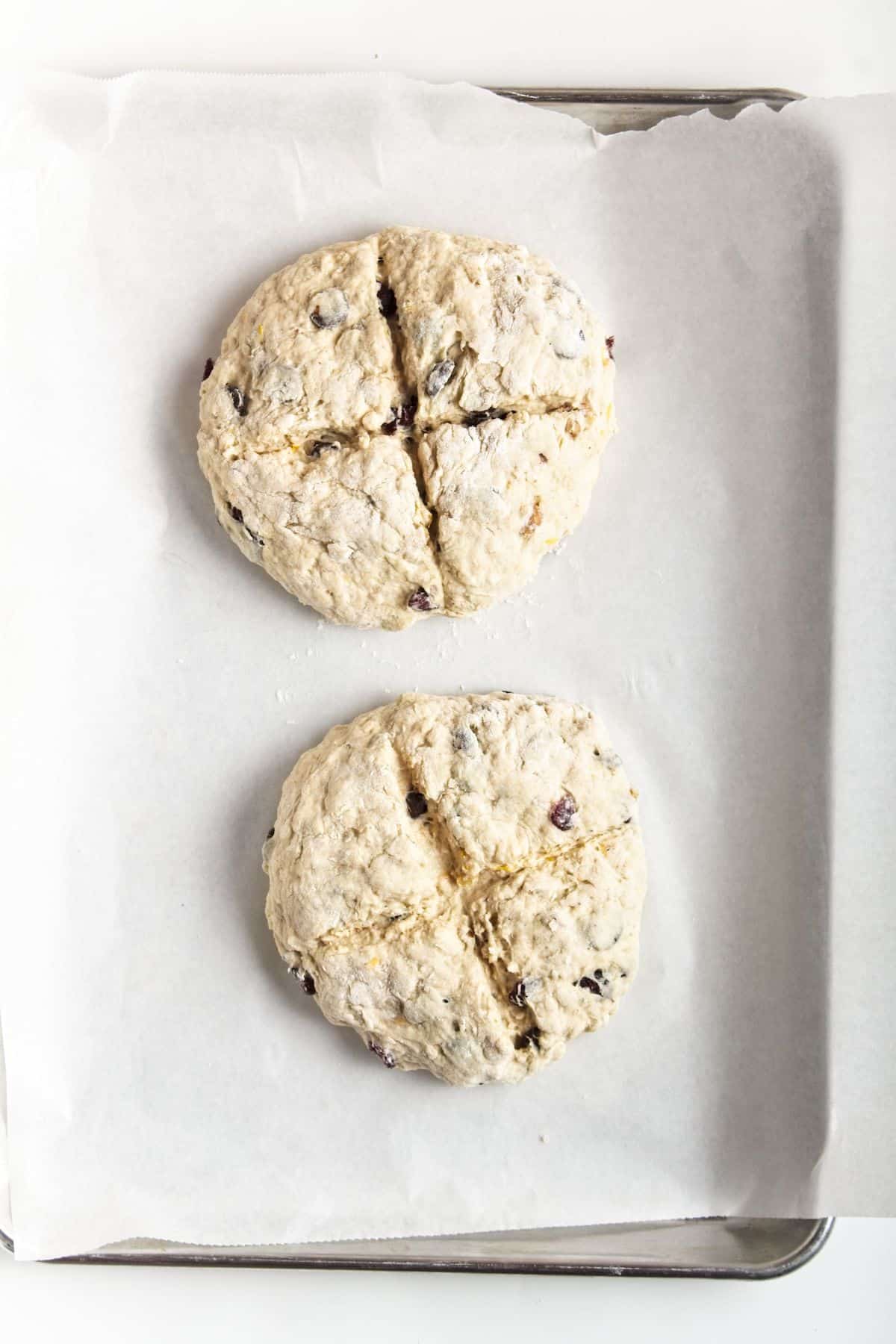 Two loaves of bread on parchment paper on baking sheet before baking.