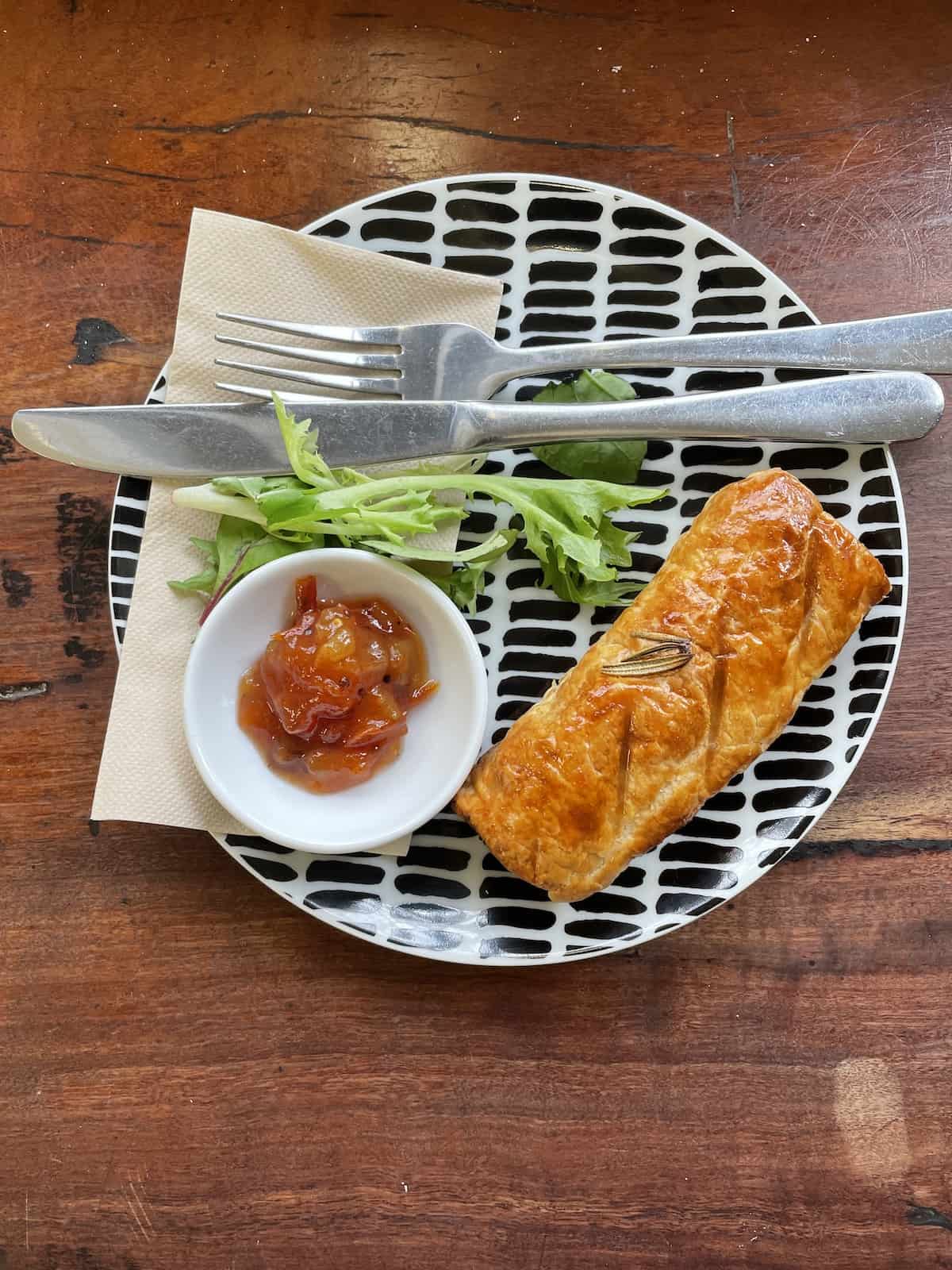 Sausage roll on a black and white plate with bowl of chutney.