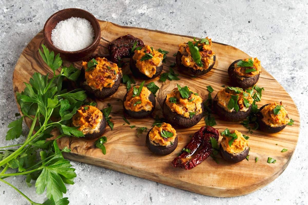 Stuffed mushrooms with slices of sundried tomato on a wood board with a bowl of salt.