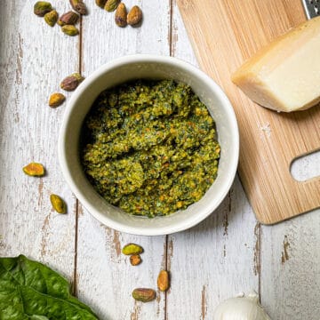 Pesto in a white bowl on a shite table with basil and a slice of Parmesan cheese on a cutting board.