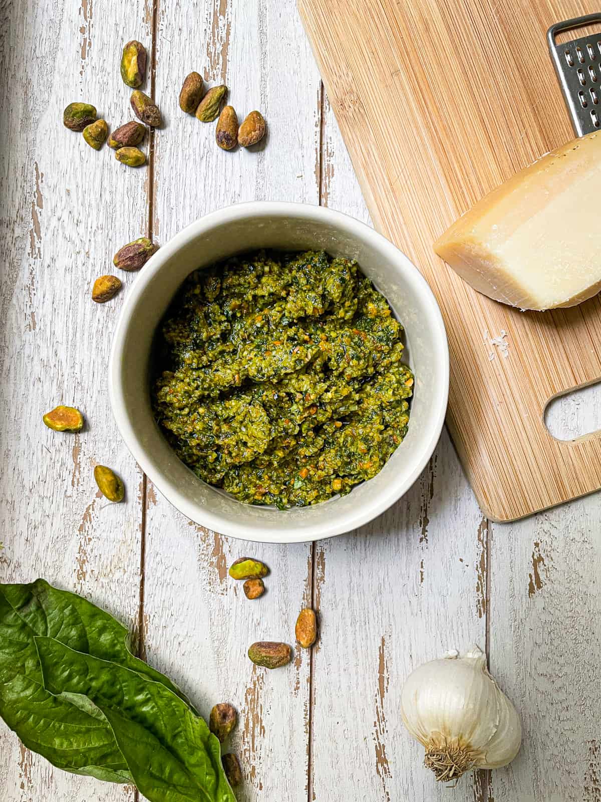 Pesto in a white bowl on a white table with basil leaves and cheese on cutting board.