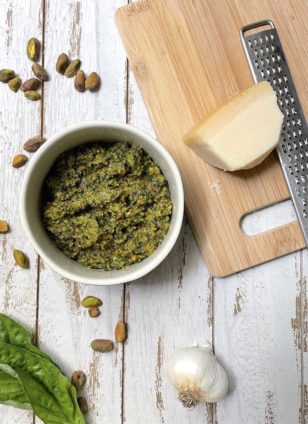 Pesto in a white bowl on a white table with basil leaves and cheese on cutting board.