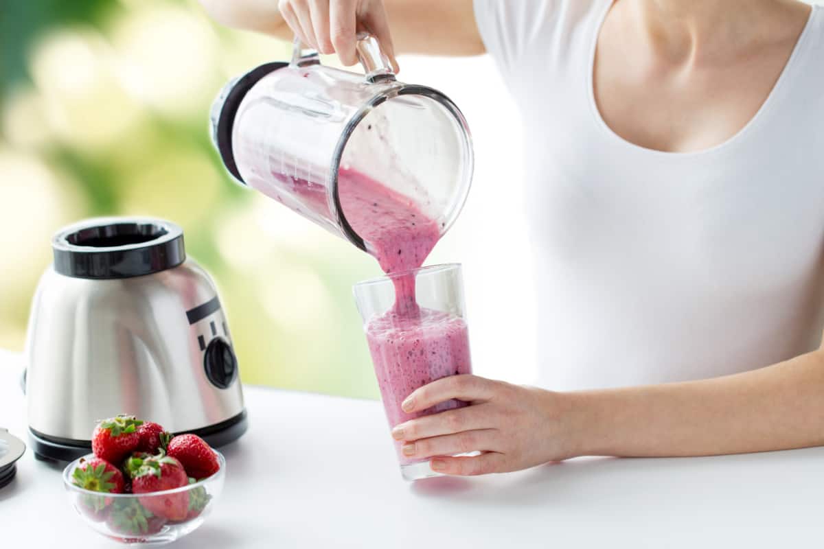 Woman holding glass of smoothie with blender and strawberries on counter.