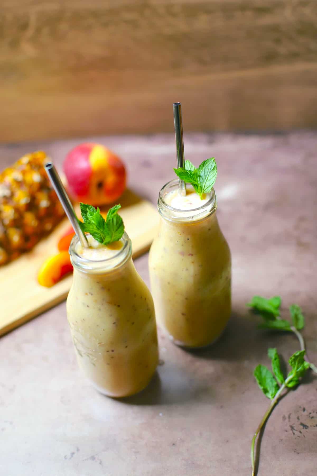 Peach smoothie in a bottle with mint and a straw with peach and pineapple in background.
