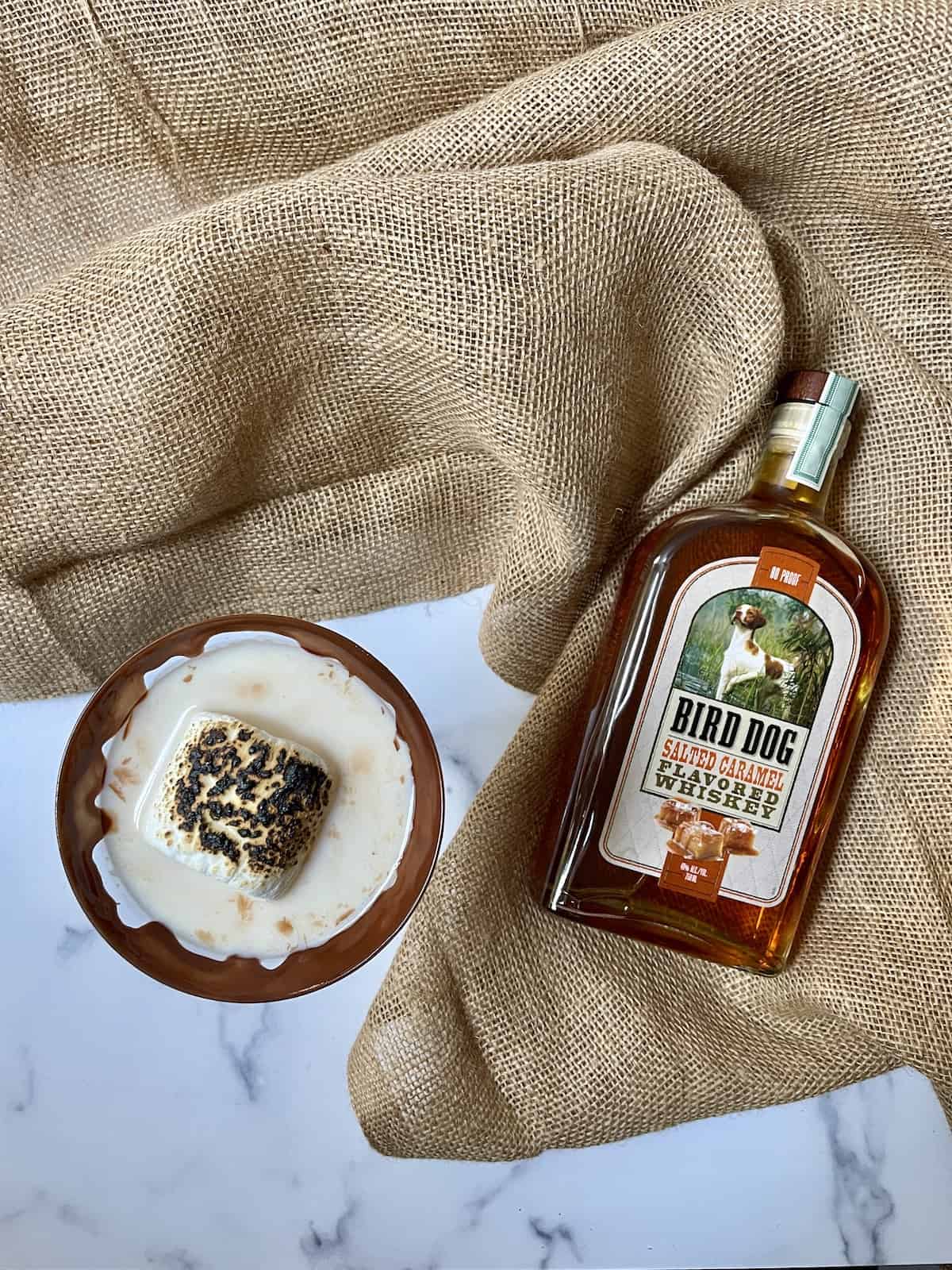 Cocktail topped with a marshmallow and a bottle of whiskey on burlap.