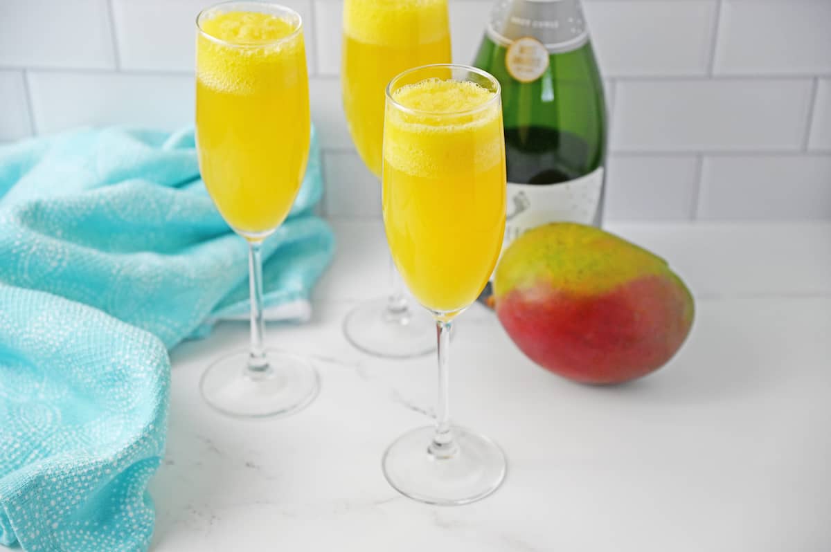 Mango mimosas, mango, bottle of champagne, and turquoise napkin on white counter.