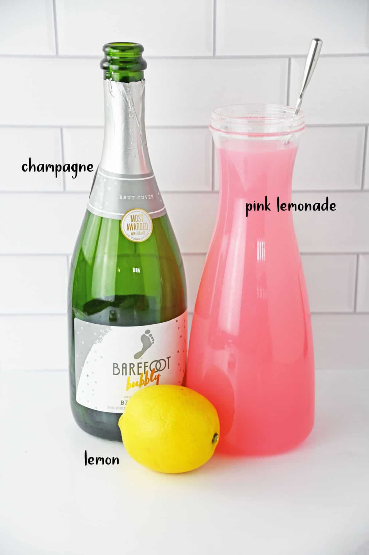 A bottle of champagne, a jug of pink lemonade, and lemons on a white counter.