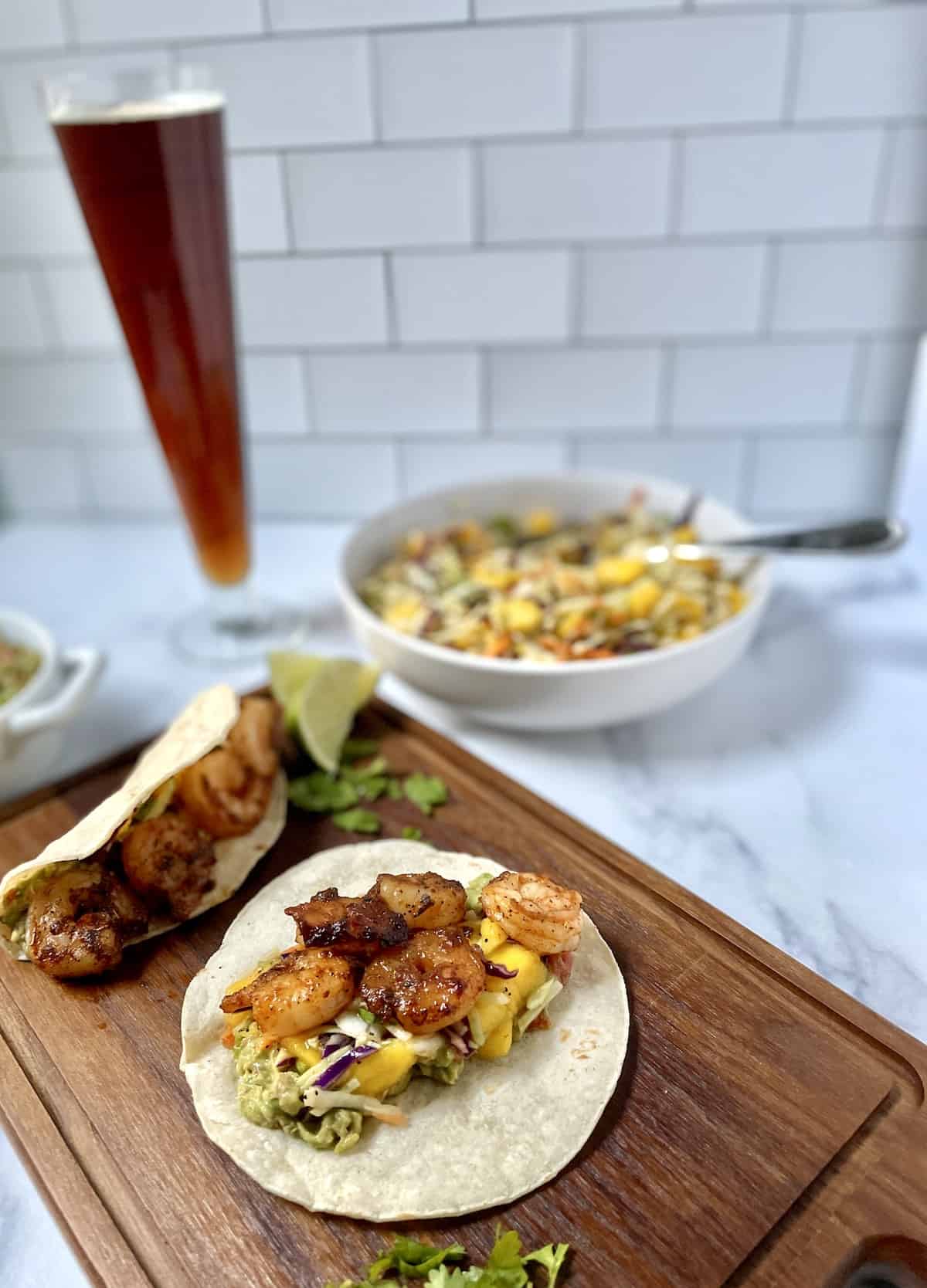 Shrimp and slaw on soft taco on wood cutting board with beer and slaw in background.