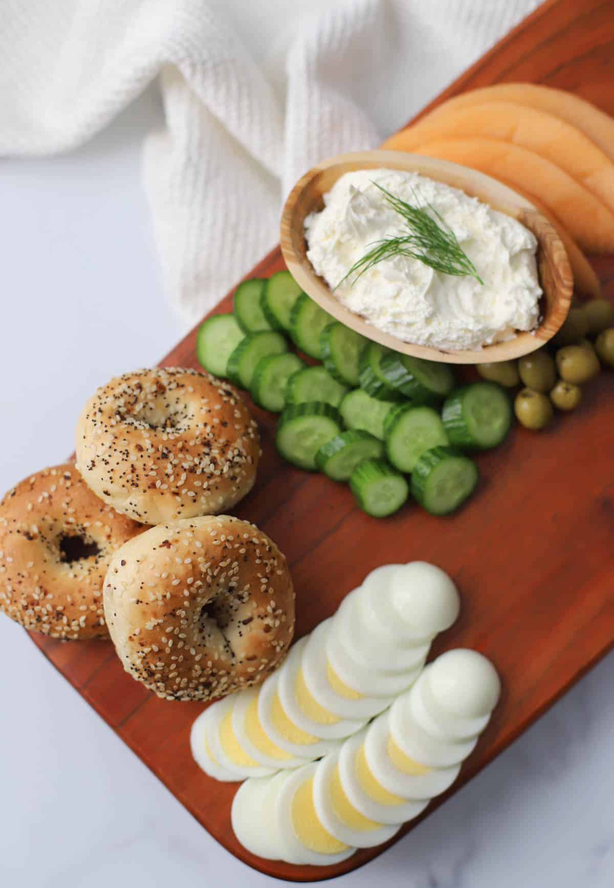 Bagels, egg, cucumber, cream cheese, olives, and cantaloupe on a wood board.