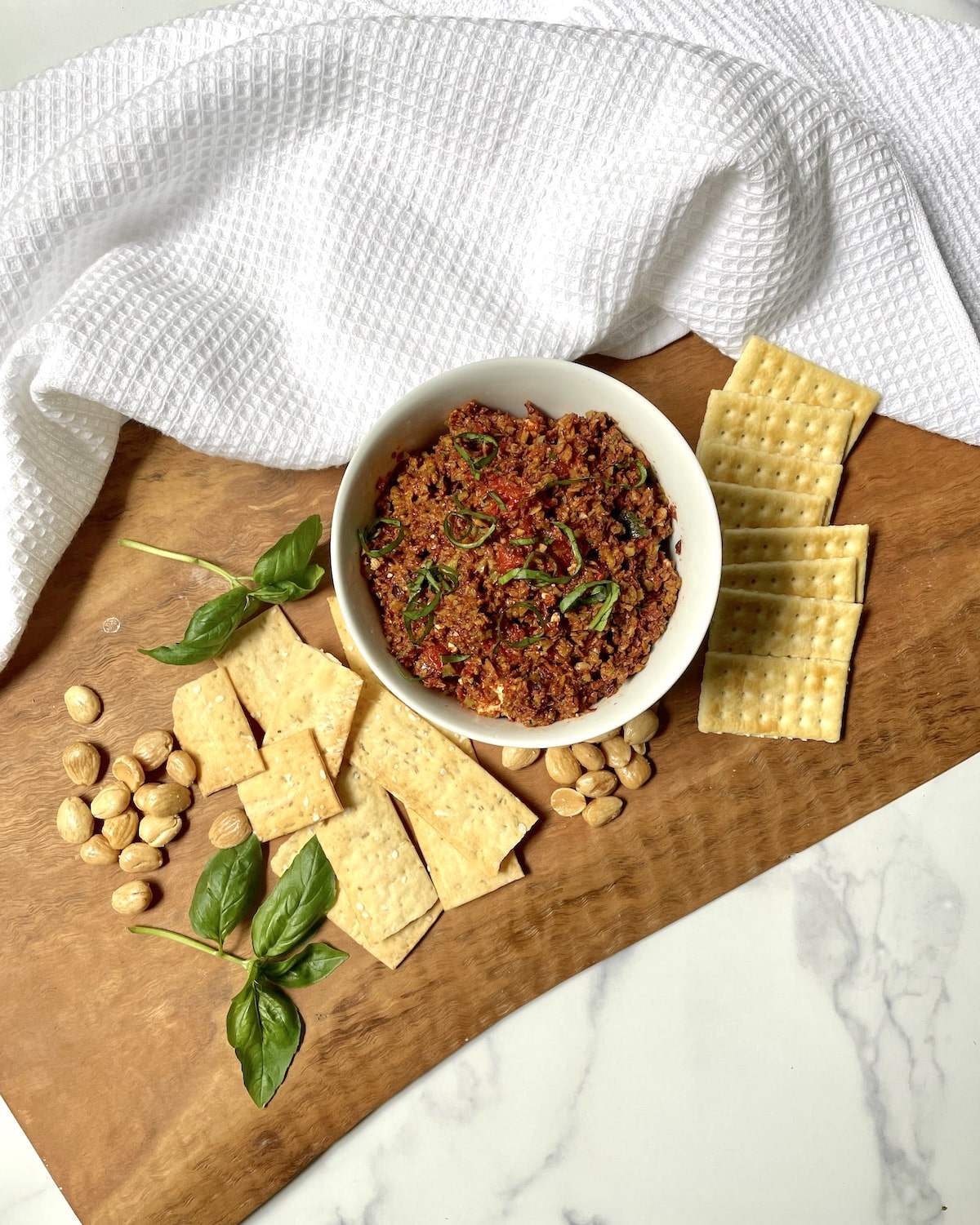 Black olive tapenade in a white bowl with crackers, nuts , and basil leaves.