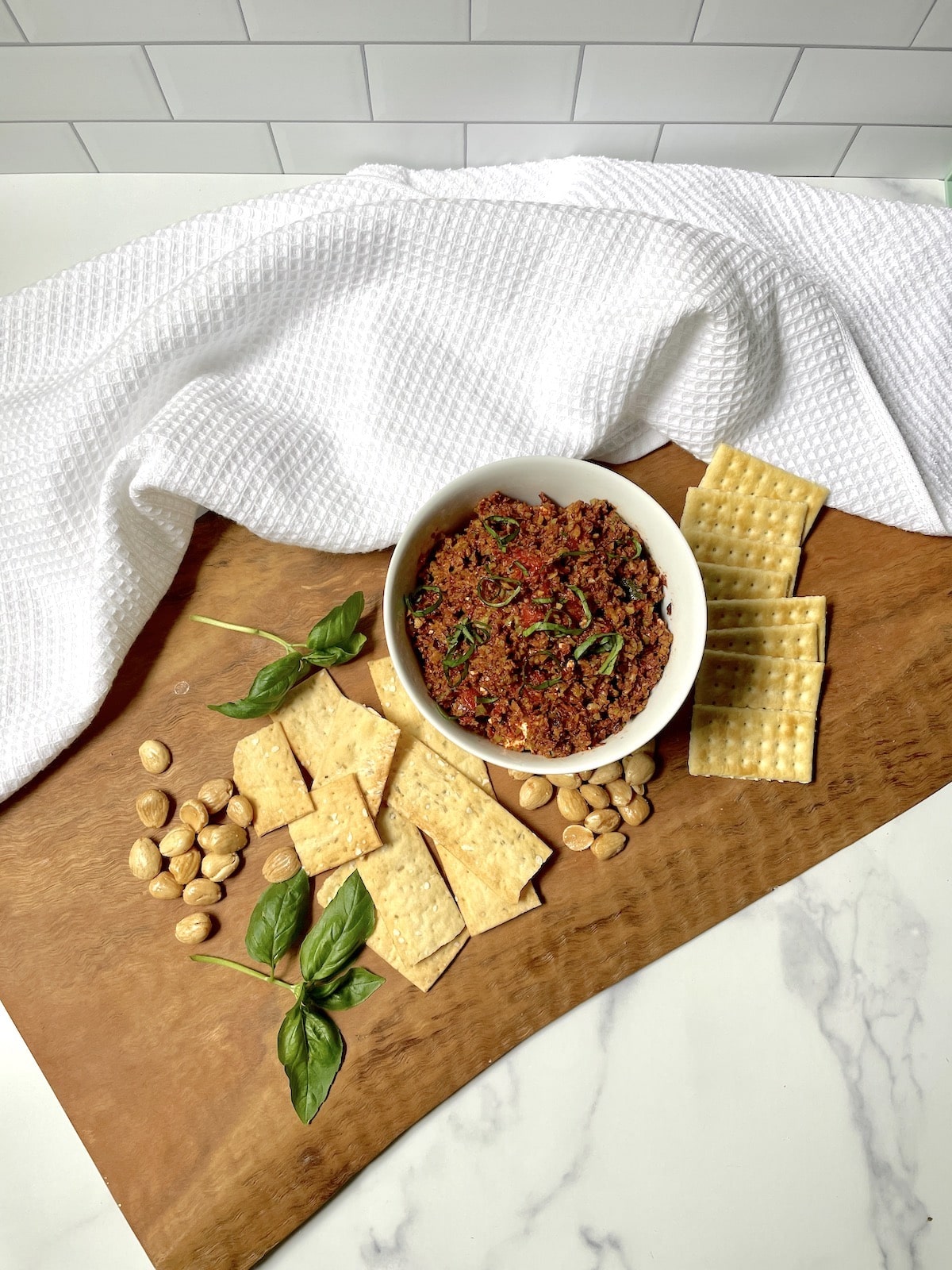 Black olive tapenade in a white bowl with crackers, nuts , and basil leaves.