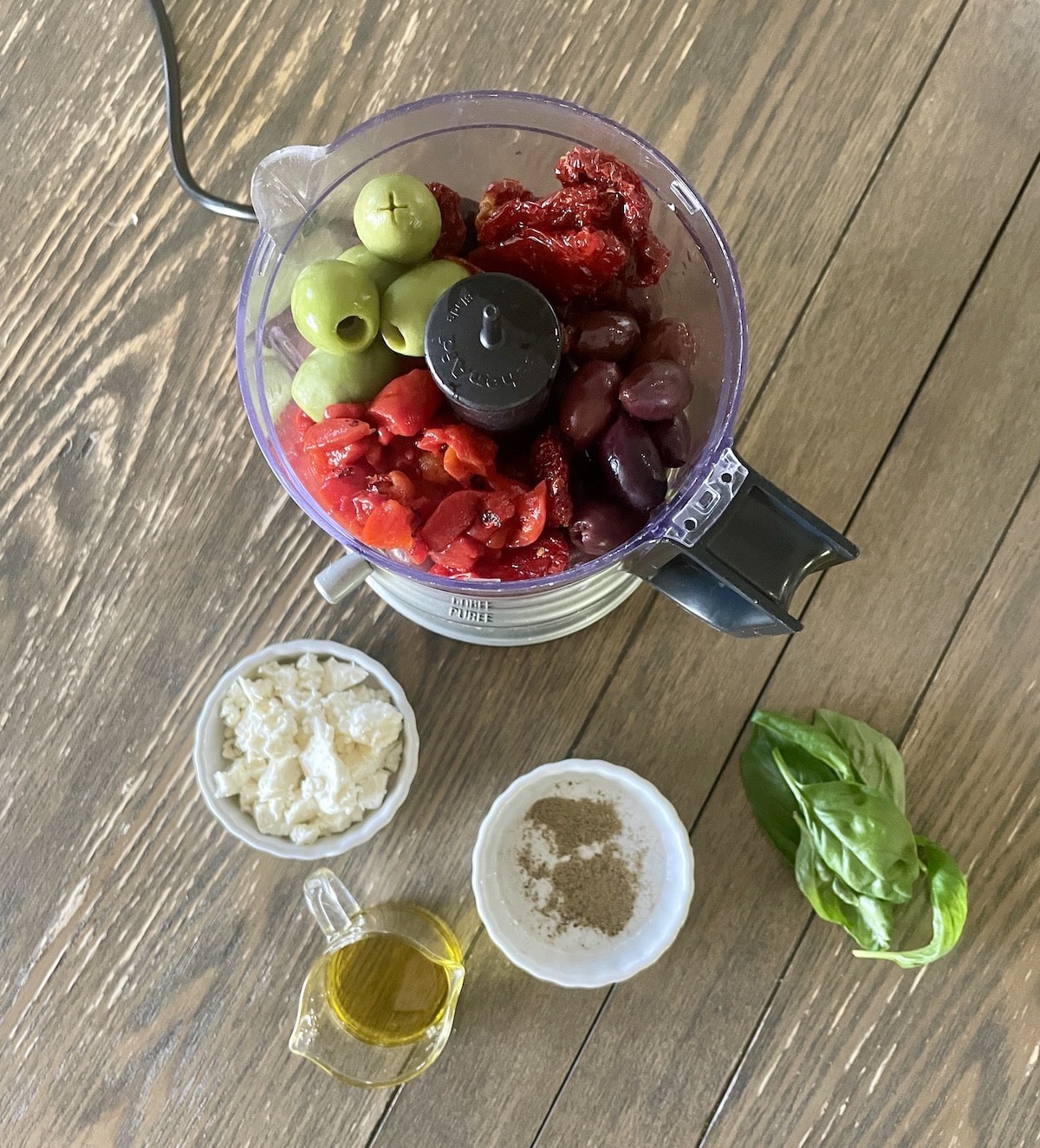 Ingredients for tapenade in a food processor with feta, pepper, olive oil, and basil on the side.