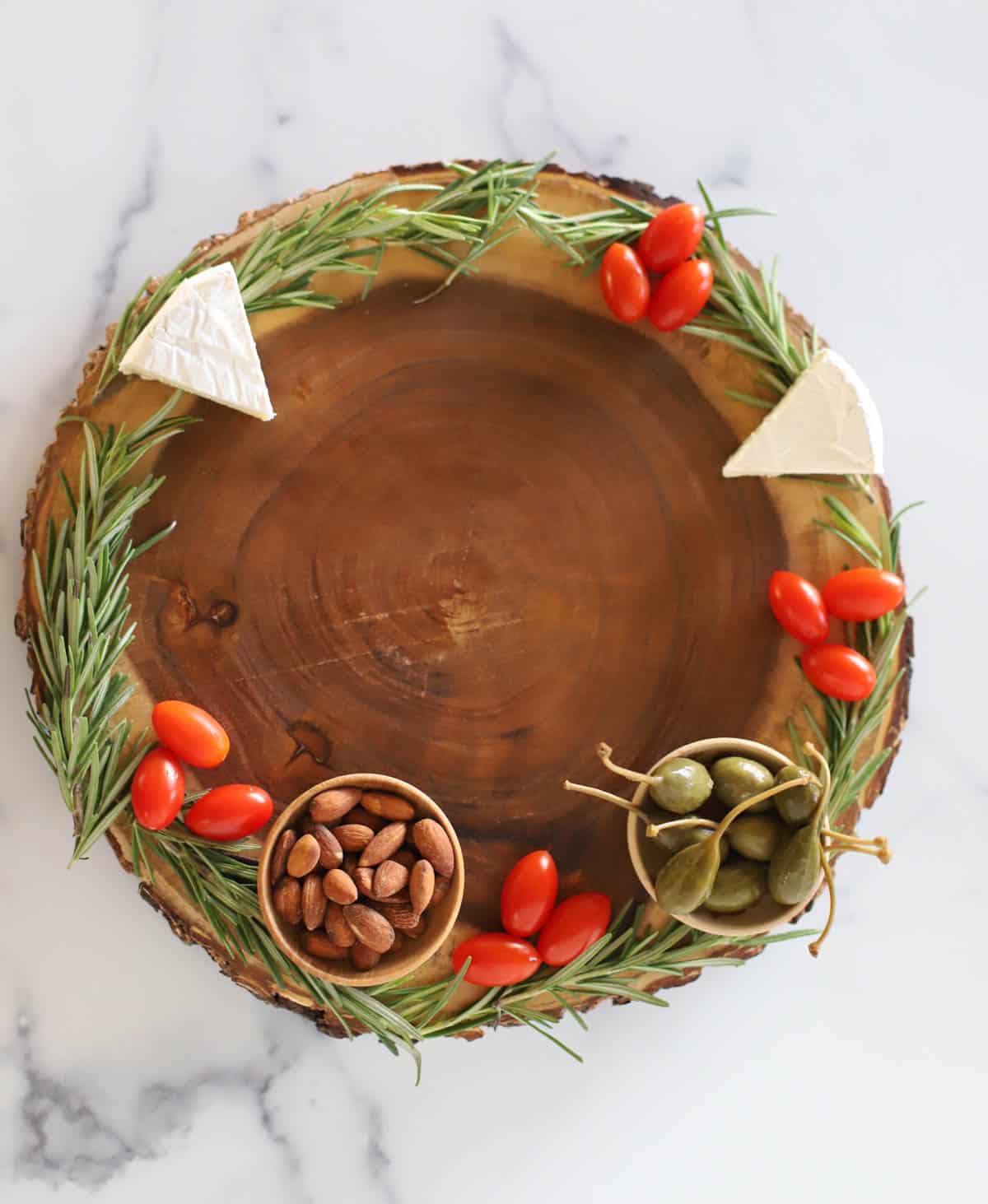 Round board trimmed with rosemary with cherry tomatoes, brie, caperberries, and almonds.