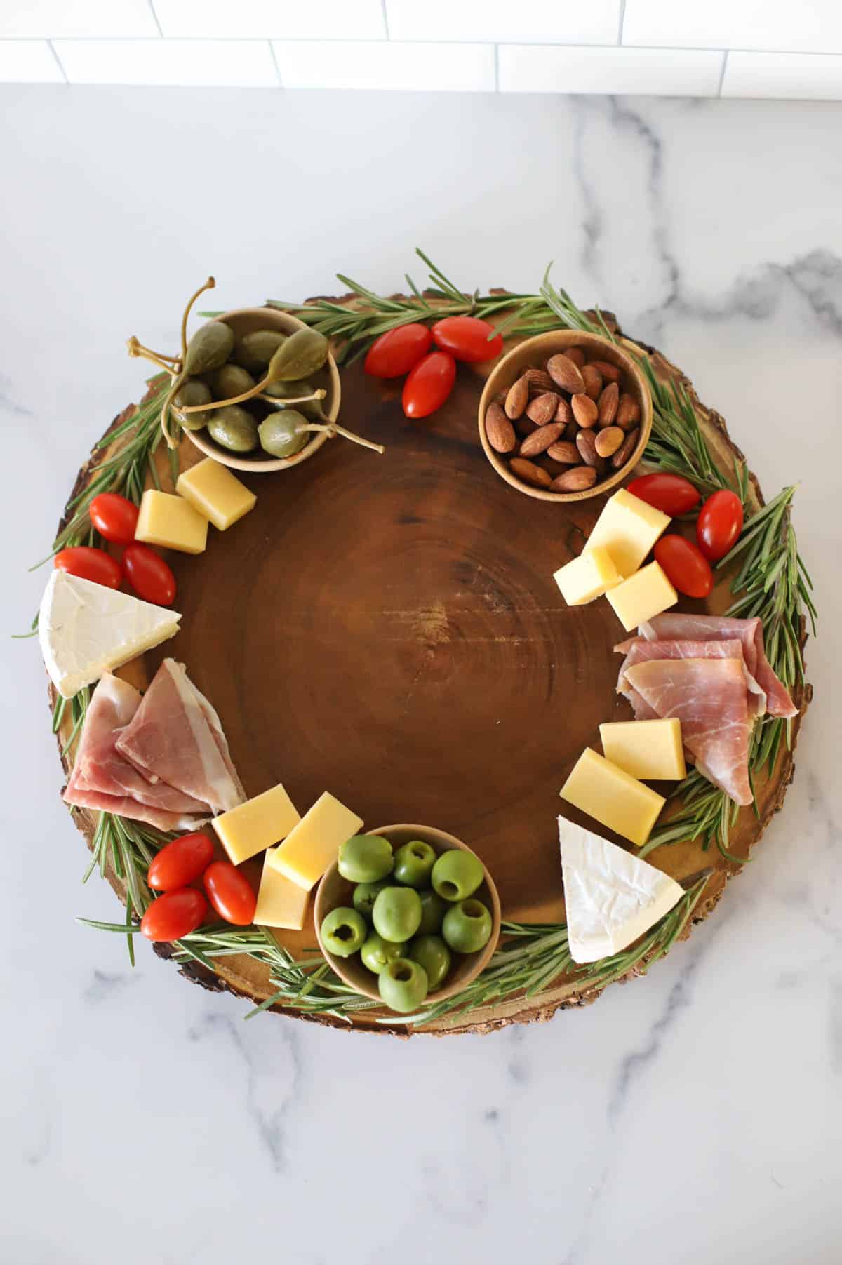 Round board trimmed with rosemary with cherry tomatoes, brie, caperberries, almonds, cheddar cheese and prosciutto.