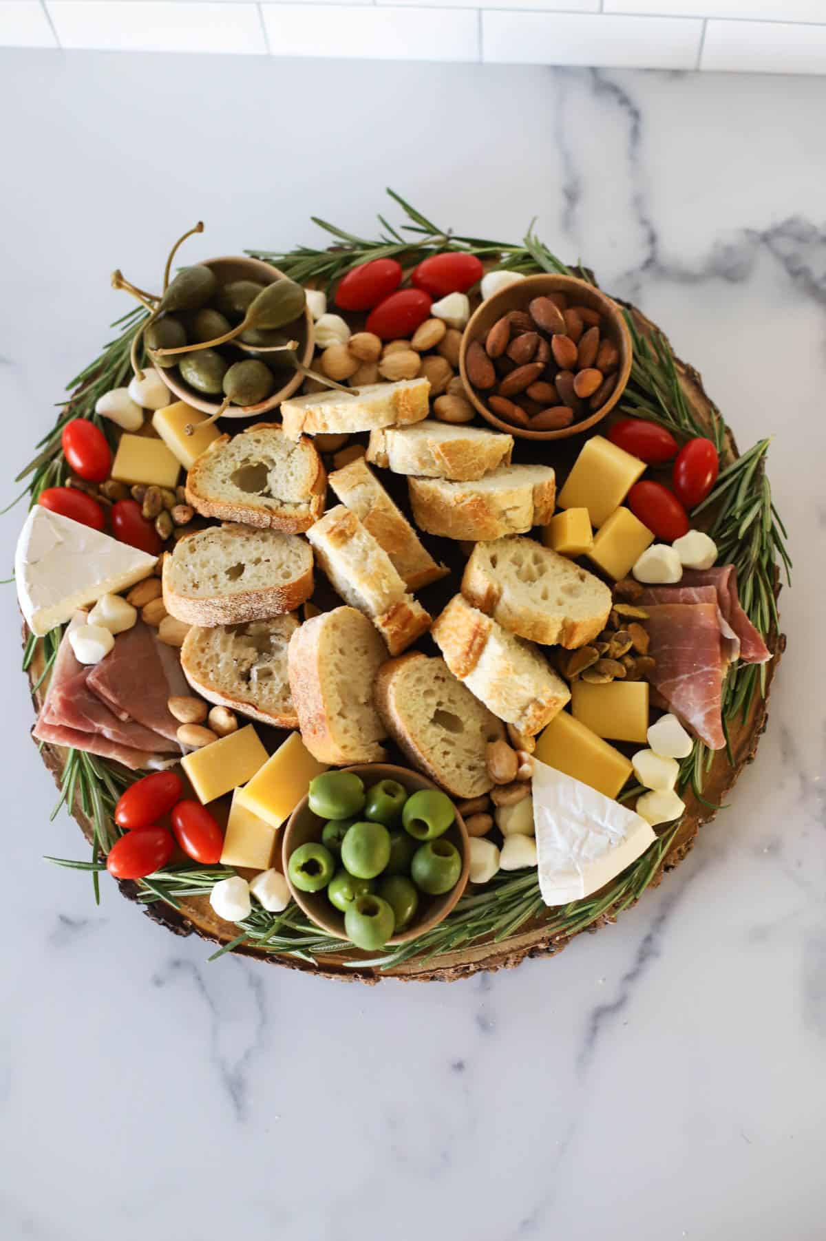 Round board trimmed with rosemary with cherry tomatoes, brie, caperberries, almonds, cheddar cheese, prosciutto, pistachios, Bucconcini, and slices of a baguette.