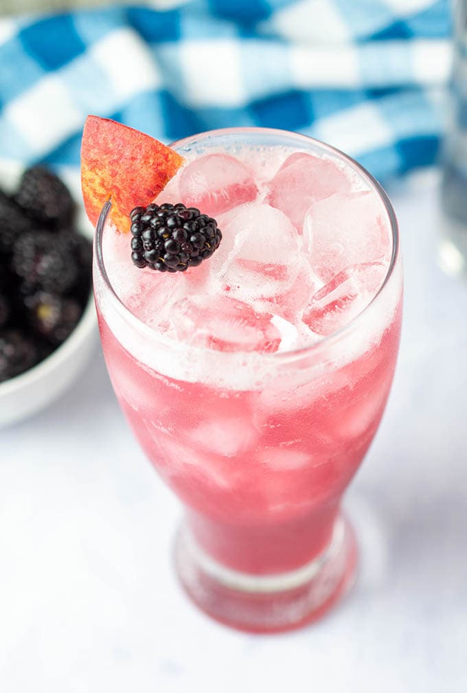 Pink-colored cocktail with a blackberry and slice of peach on top.