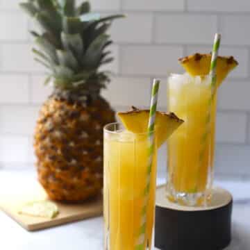 Yellow cocktail in a tall glass with pineapple wedge and straw.