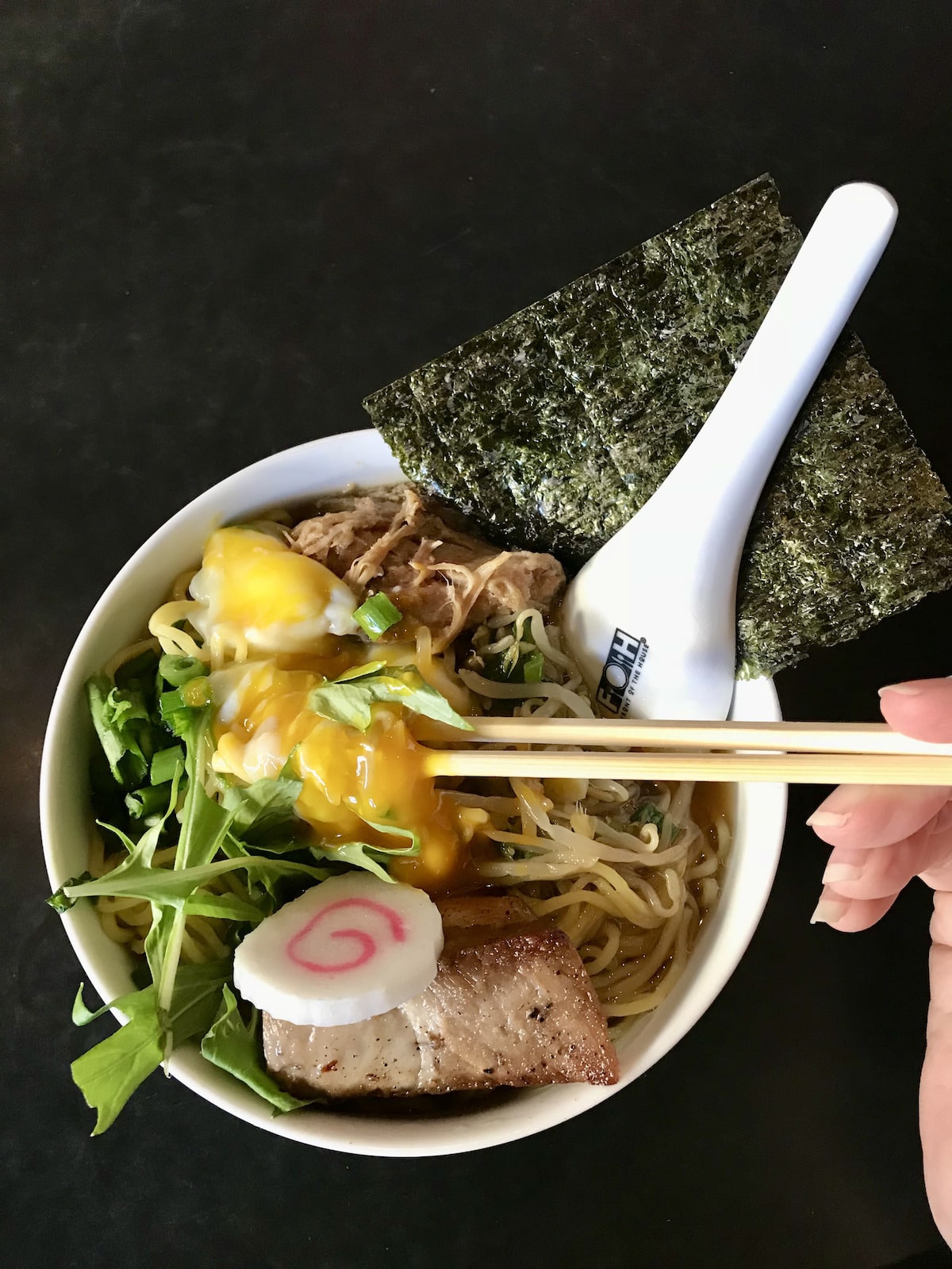 White bowl with ramen, rocket, bean sprouts, and egg with white spoon and seaweed paper.