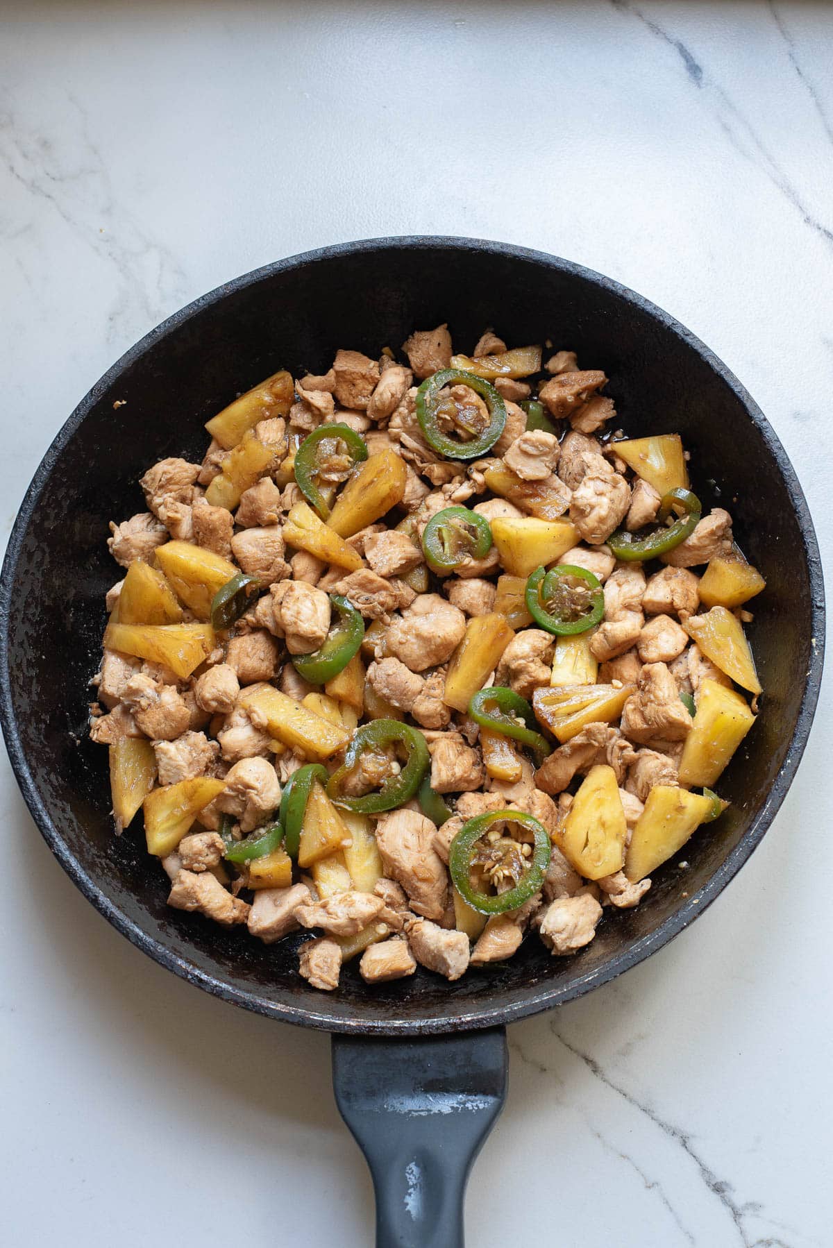 Chunks of chicken, pineapple, and jalapenos cooking in a cast iron pan.