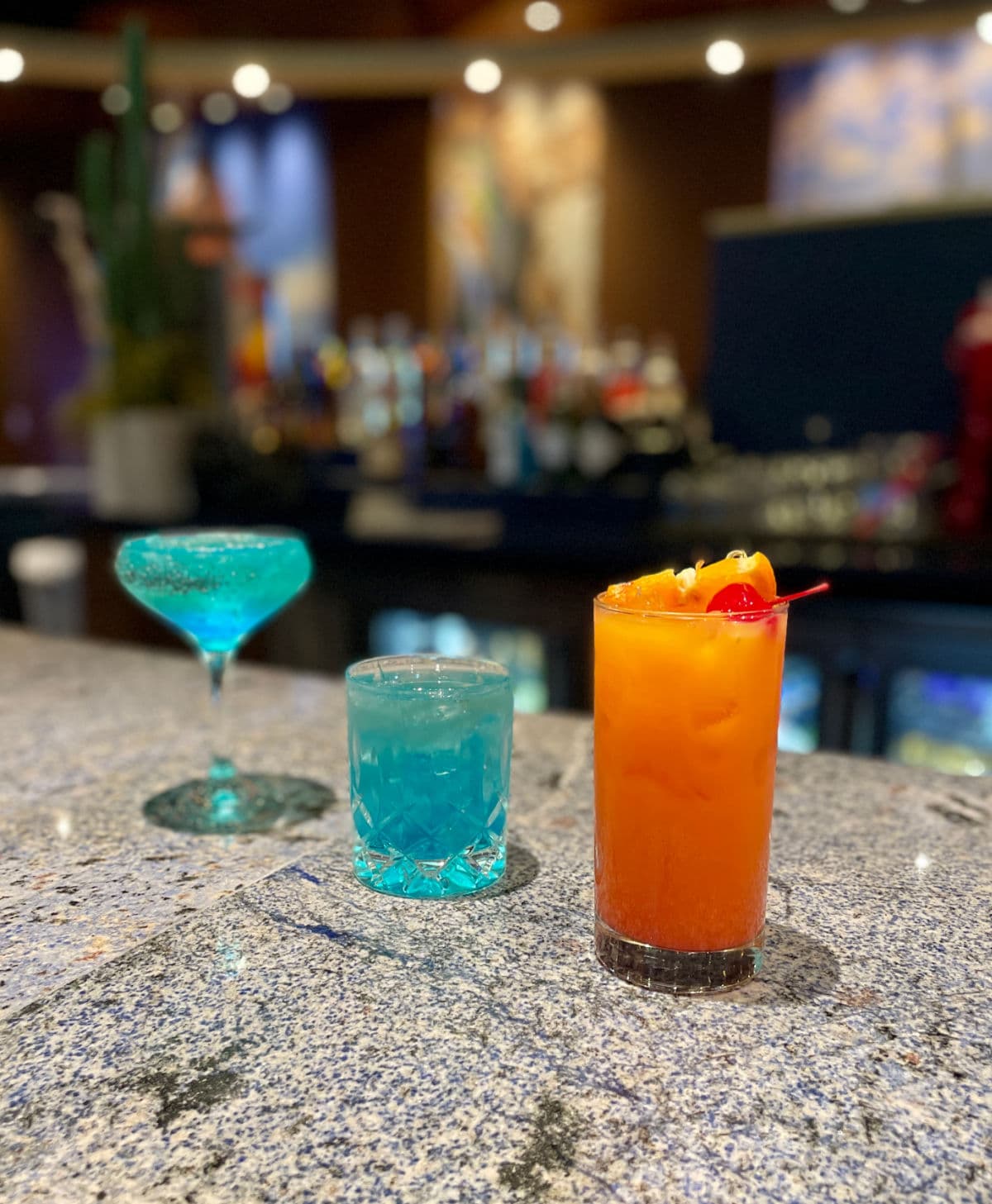 Two blue and one orange cocktail on a bar.