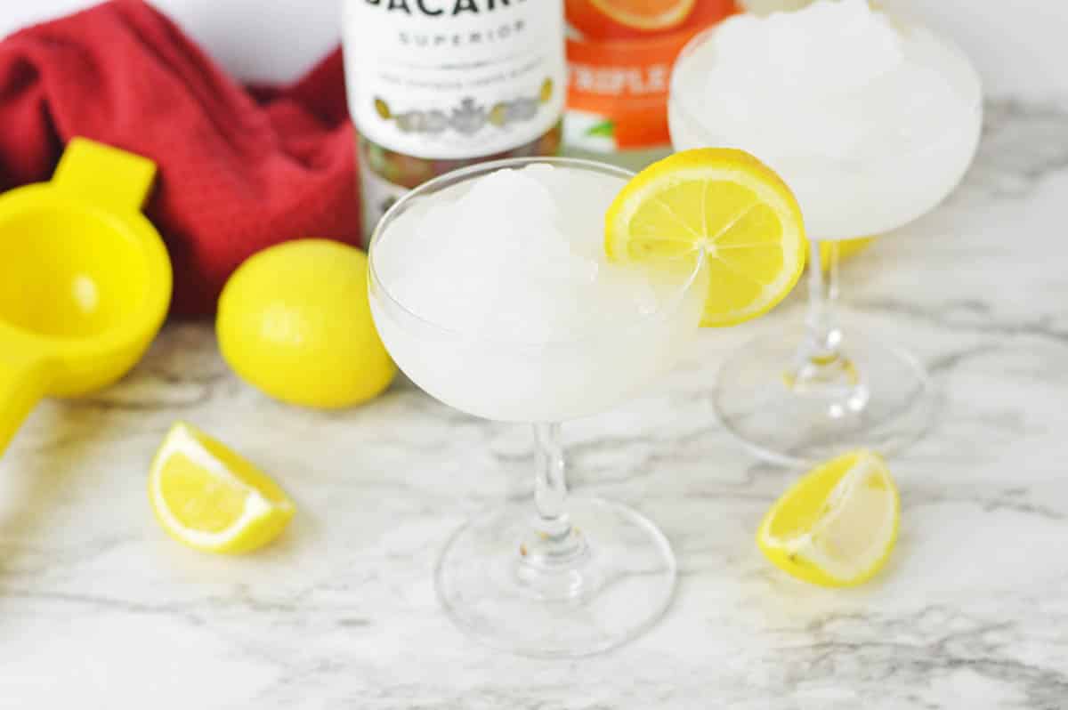 Frozen cocktail with lemon slice and lemons and bottle of rum in background.