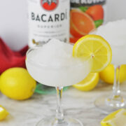 Frozen daiquiri with a lemon slice on side and lemons and bottles of alcohol in background.