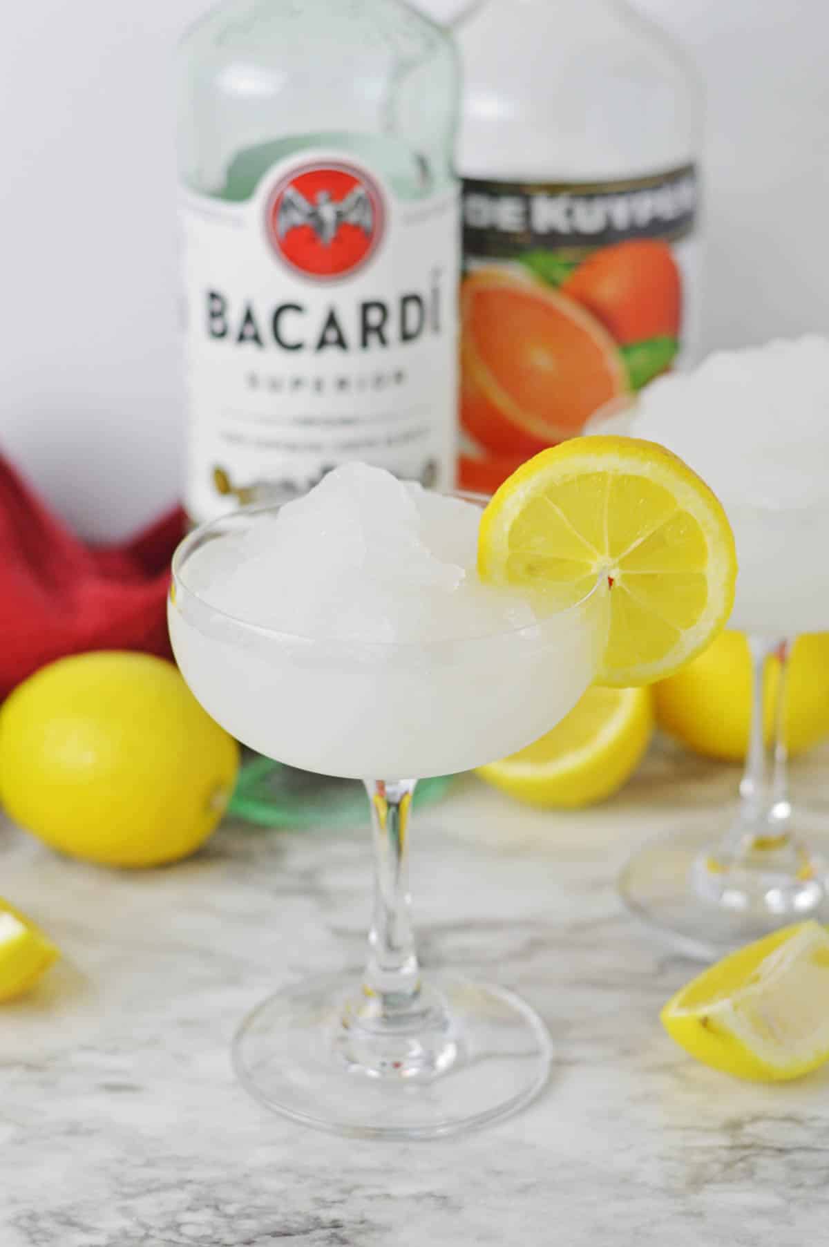 Frozen cocktail with lemon slice and lemons and bottle of rum in background.