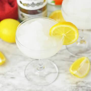 Frozen daiquiri with a lemon slice on side and lemons and bottles of alcohol in background.
