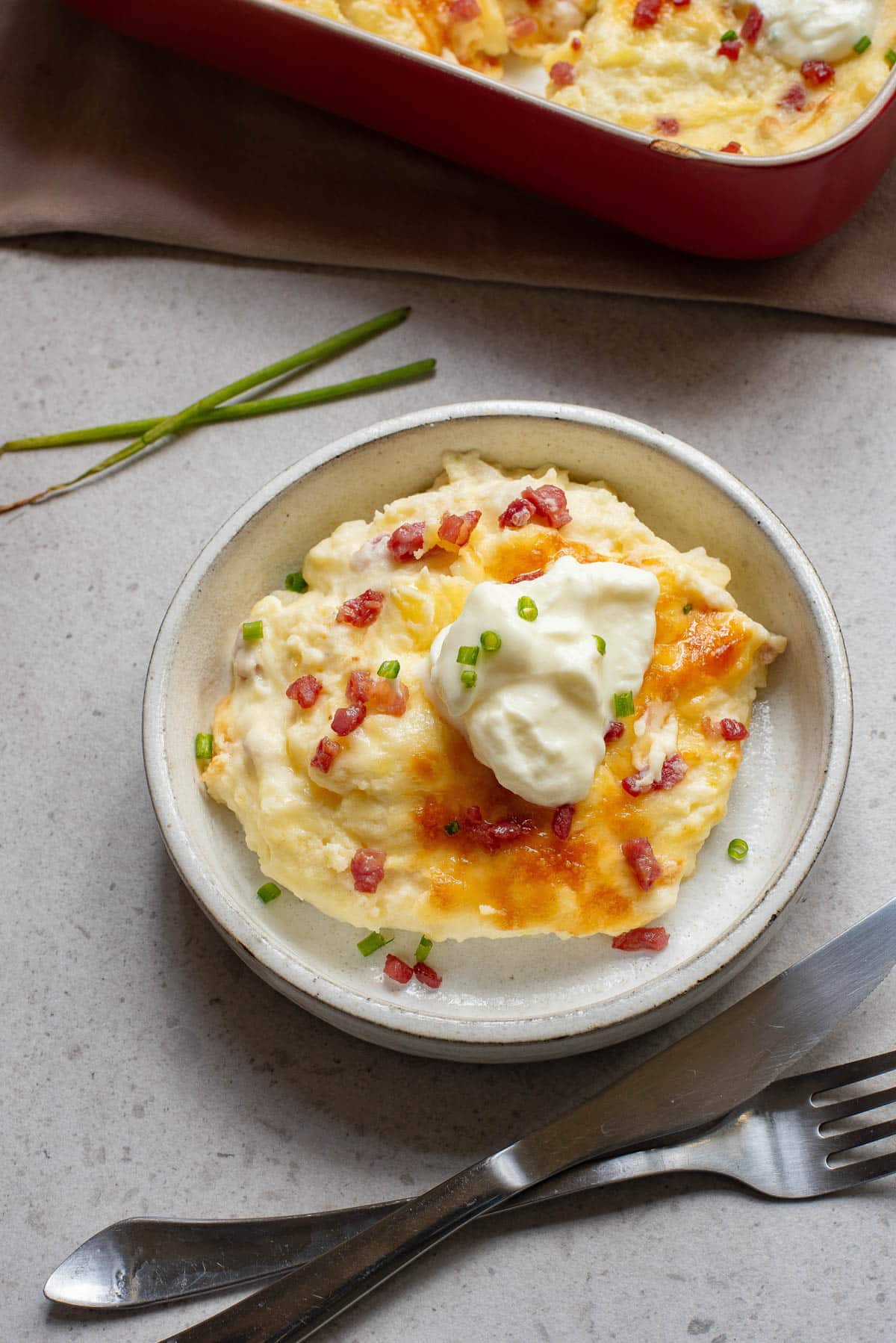 One serving of potato casserole with cheese and bacon in a white bowl with a knife and fork.