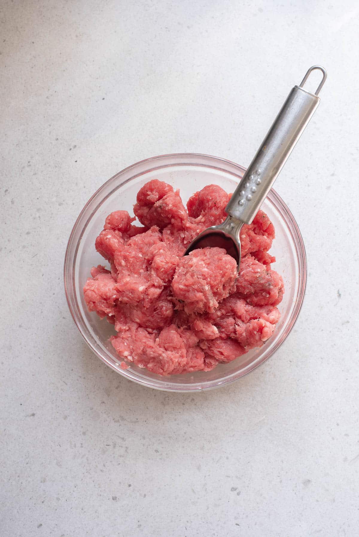 Raw ground beef meatballs in a glass bowl with scoop.