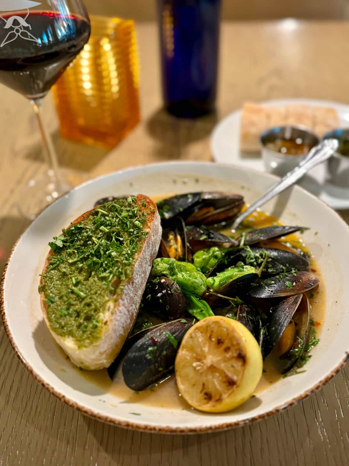 Mussels with bread and lemon half in a white bowl with glass of wine in background.
