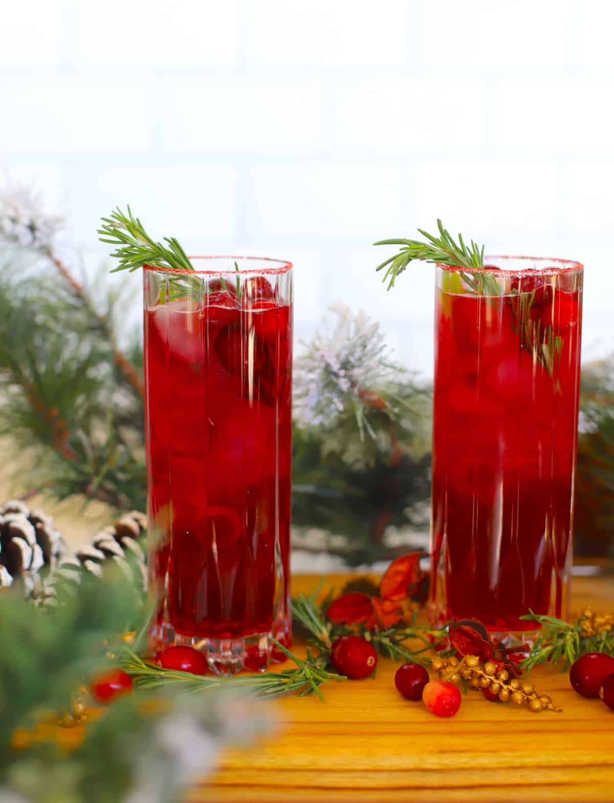 Tall glasses filled with cranberry juice and fresh berries and rosemary with holiday decor.