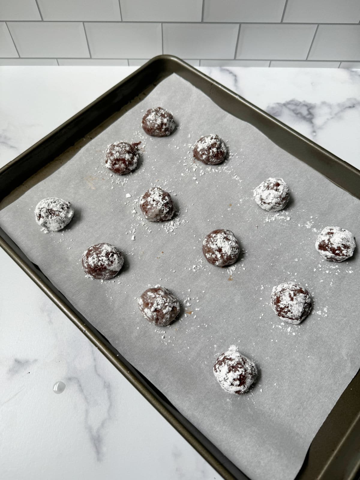 Chocolate dough balls covered in powdered sugar on parchment on baking tray.
