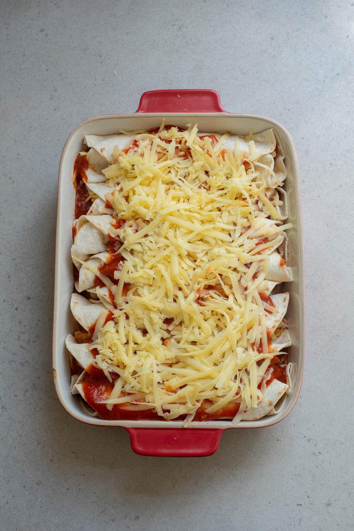 Enchiladas in a red baking dish covered with shredded cheese before baking.