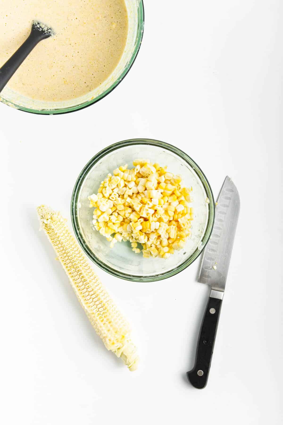 Cornbread batter in a glass bowl and a small glass bowl with corn kernels and empty cobs.