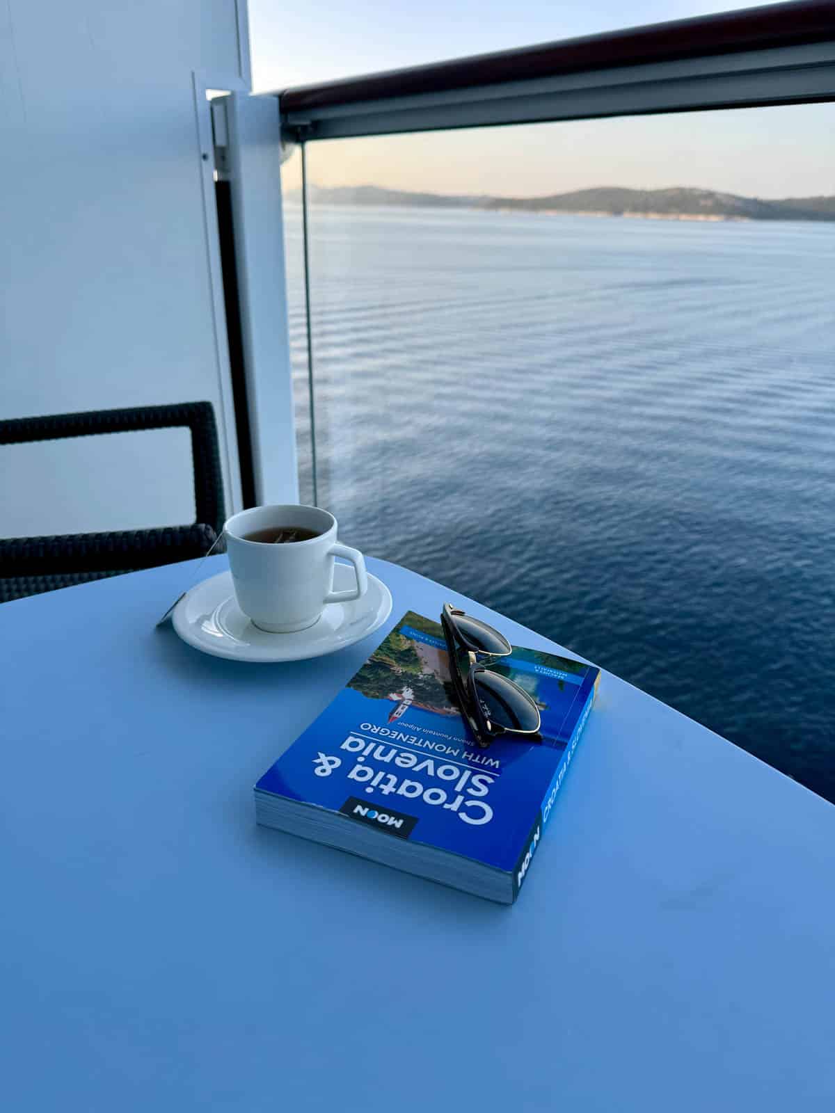 Table on the balcony of a cruise ship with cup of tea, book, and sunglasses.
