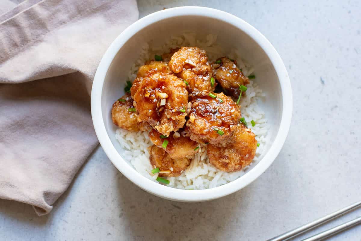 Fried shrimp with sauce and sesame seeds and chives in small white bowl with chopsticks and naplkin.