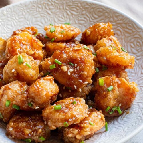 Fried shrimp with sauce and sesame seeds and chives in white bowl with chopsticks.