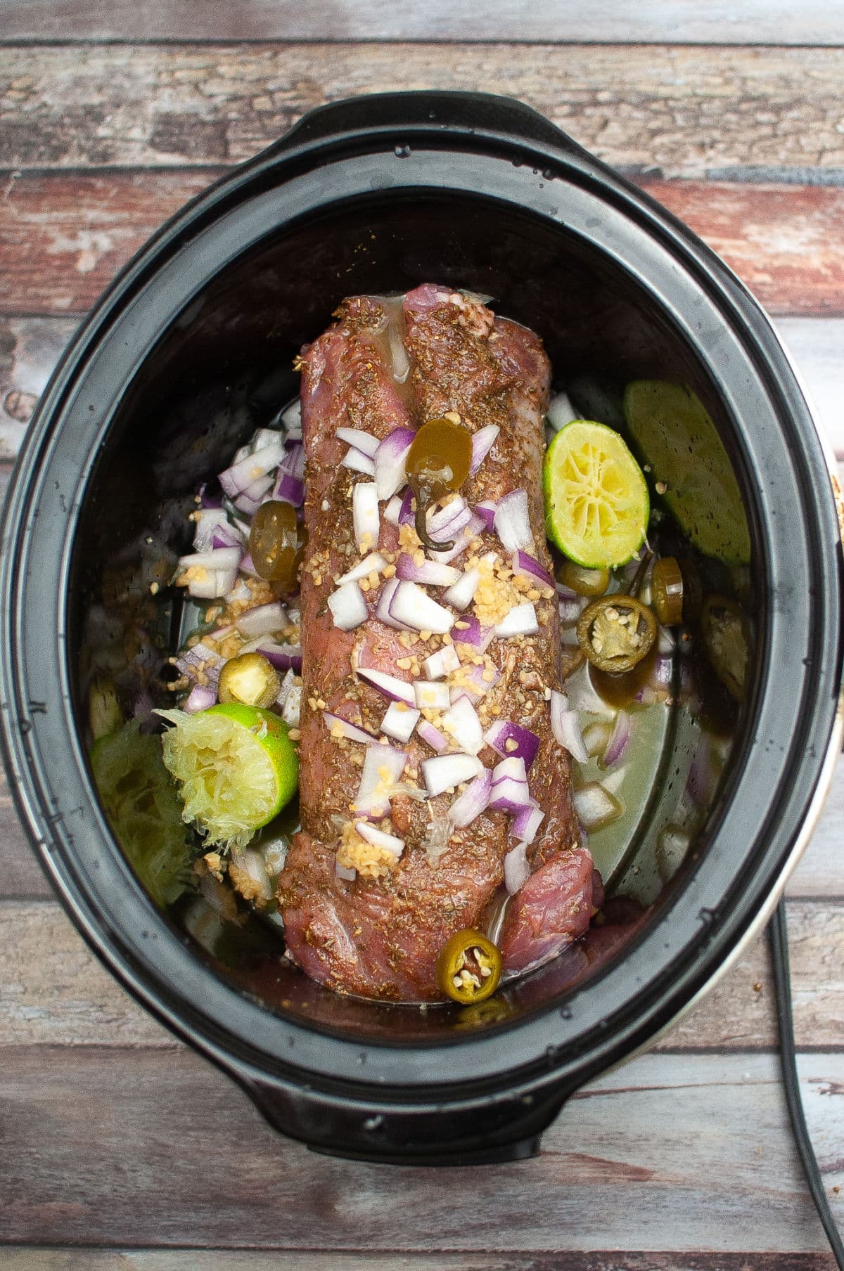 Uncooked pork tenderloin with onions, jalapeños, spices and limes in juices in slow cooker.