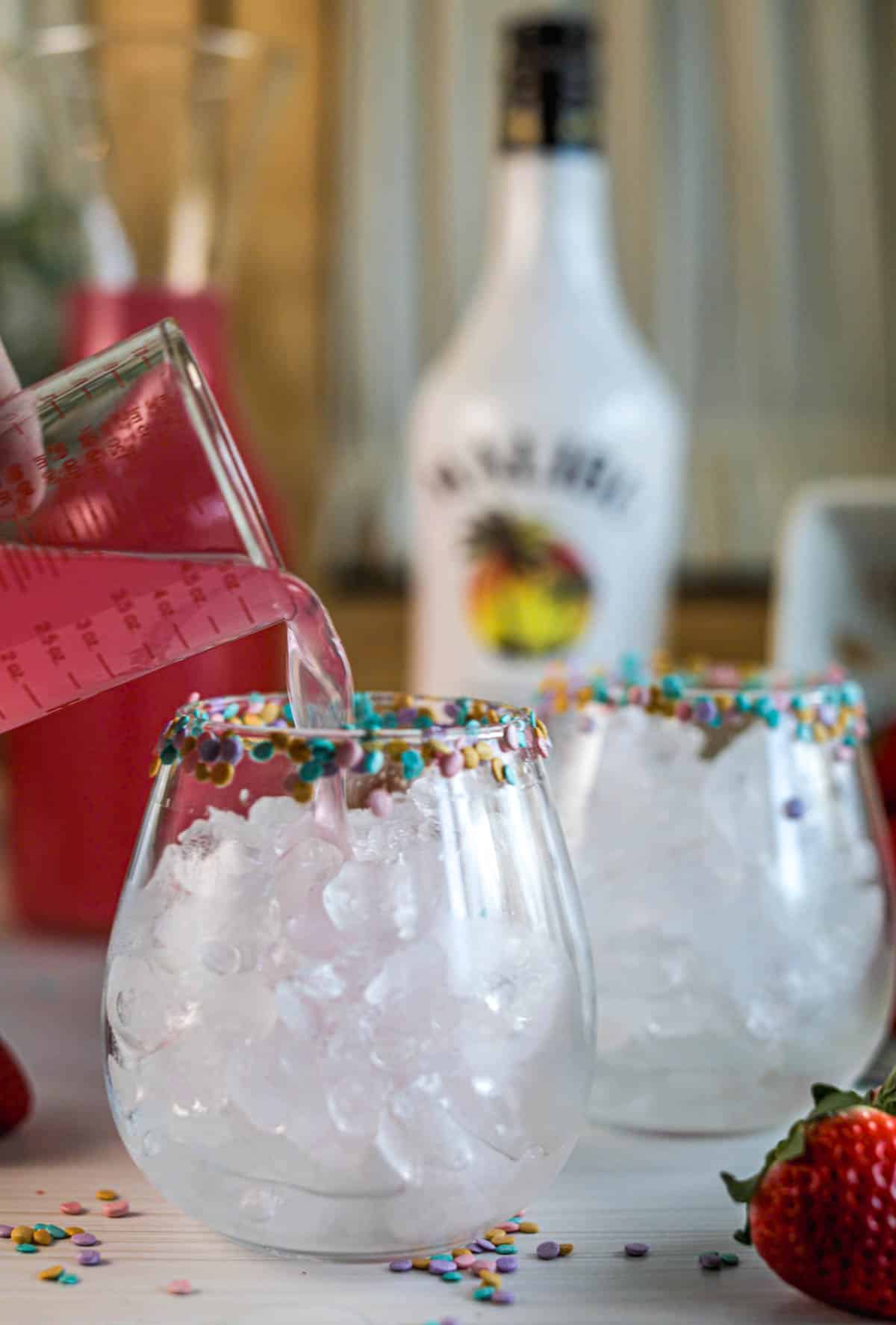 Pouring pink lemonade into glasses of ice with sprinkles on rim.