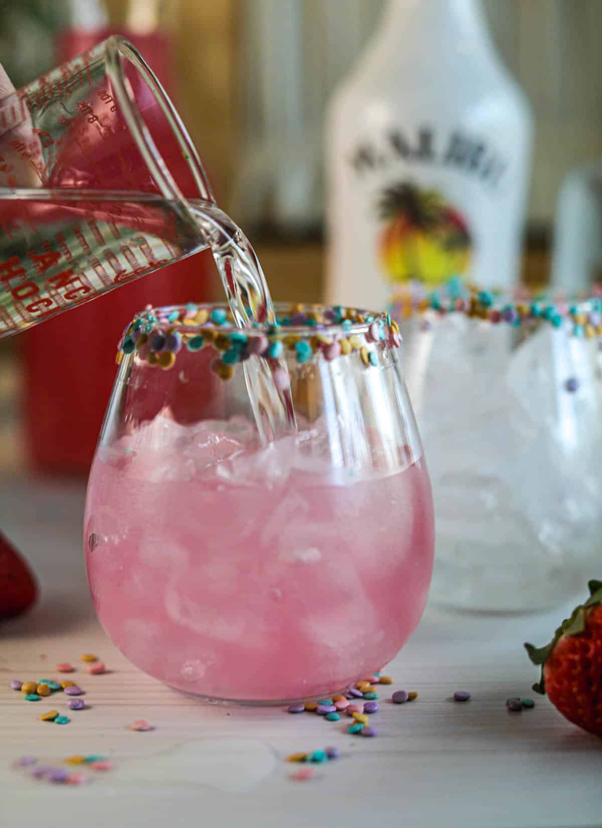 Adding rum into glasses of pink lemonade with sprinkles on rim.