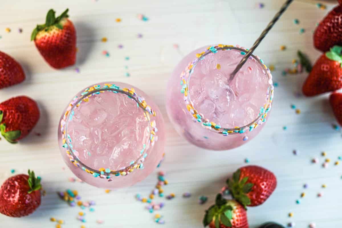 Pink cocktails with sprinkles on rim and stir stick with strawberries and sprinkles on table.