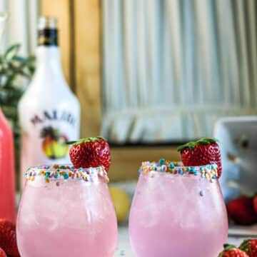 Pink cocktails with sprinkles on rim with strawberries and sprinkles on table.
